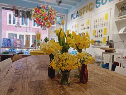 Narcissen van verschillende groottes in verschillende vaasjes op een houten tafel in een kleurrijke winkel. Op de muur staat plastic soep geschreven
