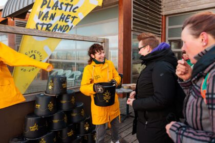 Lachende dame in contact met 2 andere dames, biedt hen een emmer aan. op vlag staat te lezen, voeten in het zand, plastic van het strand