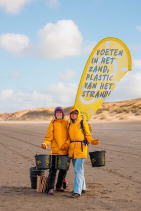 2 lachende dames in gele regenjassen op het strand met emmers in de handen en een vlag met de tekst voeten in het zand, plastic van het strand
