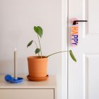 Roze deurhanger met het woord happy en sad hangend aan binnendeur naast een dressoir met plant en kandelaar.