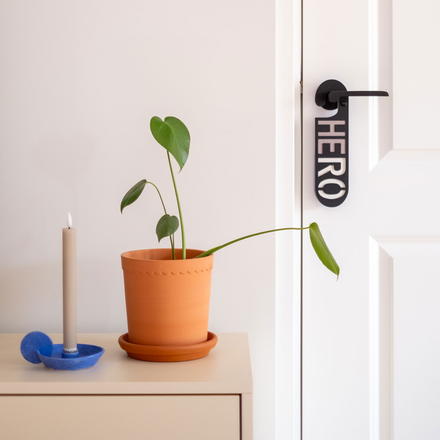 Zwarte deurhanger met het woord hero hangend aan binnendeur naast een dressoir met plant en kandelaar.