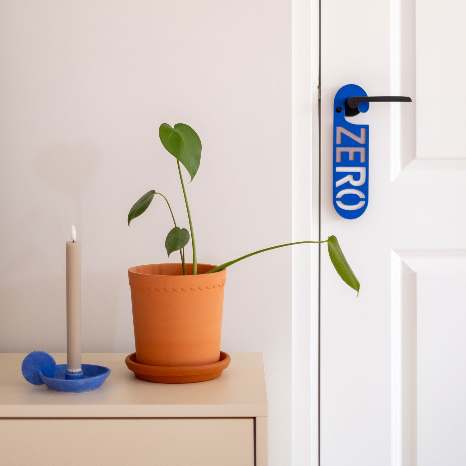 Blauwe deurhanger met het woord zero hangend aan binnendeur naast een dressoir met plant en kandelaar.
